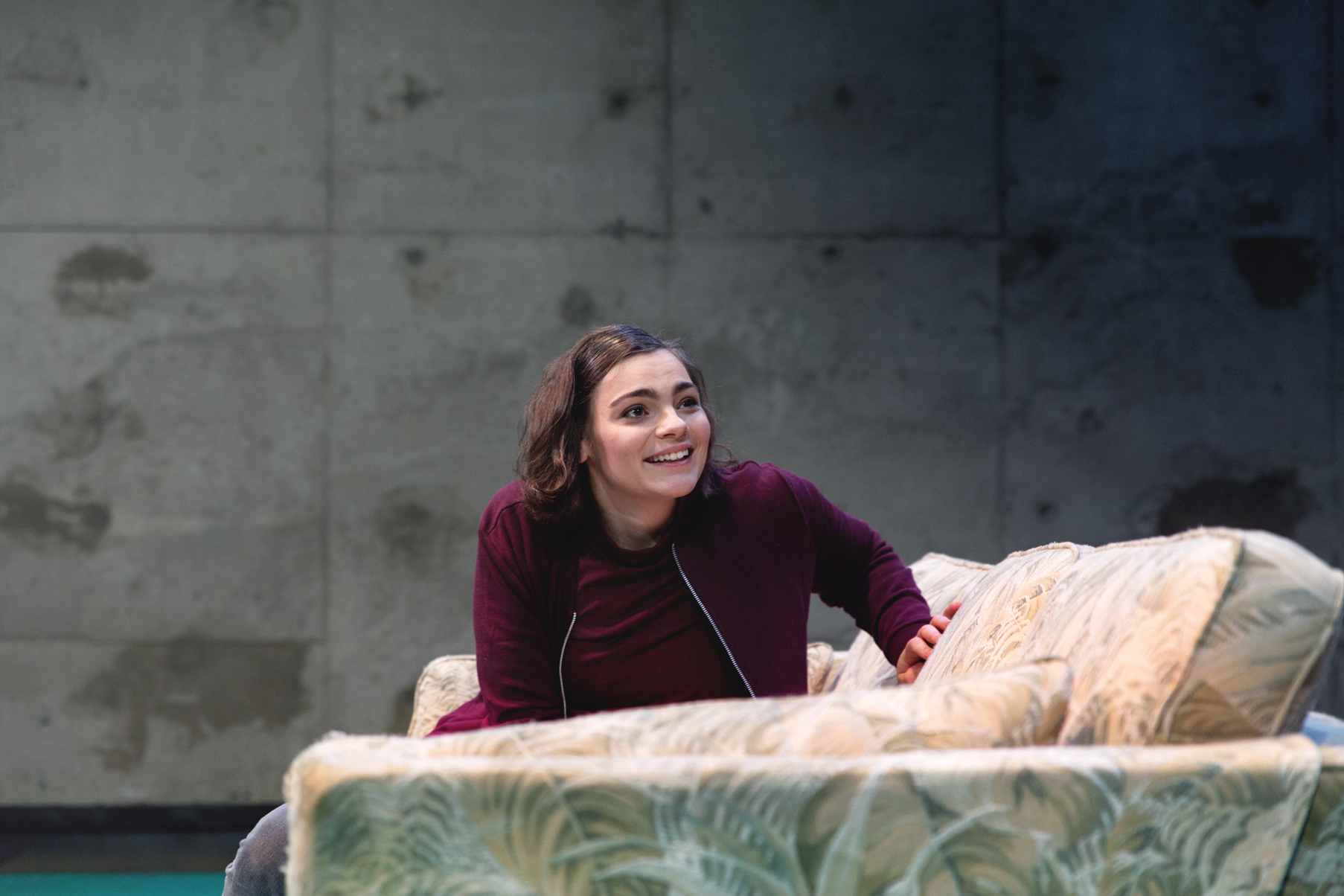 Linnea Sundblom sitter leende vid en säng på en teaterscen.