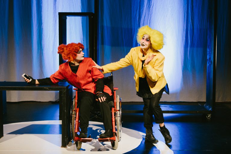 Två skådespelare i färgglada kläder – en i rullstol – i livlig scen.